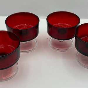 Puede incluir: Cuatro copas de postre de vidrio rojo rubí con tallos de vidrio transparente. Los cuencos tienen forma redondeada y están sobre una superficie blanca. El vidrio tiene un acabado brillante.