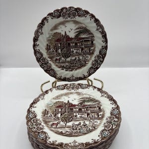 Op de afbeelding: Een stapel vintage bruin-witte borden met een gedetailleerde landschapsafbeelding. De borden hebben een geschulpte rand en een decoratieve rand. Eén bord wordt op een goudkleurige standaard getoond.