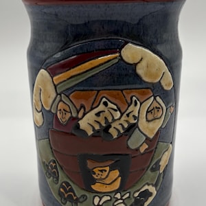 Könnte beinhalten: Eine blaue Keramiktasse mit rotem Rand, die eine handgemalte Szene von Noahs Arche mit Tieren und einem Regenbogen zeigt. Die Arche ist braun mit schwarzem Innenraum, und die Tiere sind weiß, braun und schwarz. Der Hintergrund ist blau mit weißen Wolken.