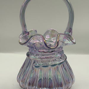 Peut inclure: Panier en verre irisé avec un bord à volants et une anse incurvée. Le panier a une base bulbeuse avec des rainures verticales et une spirale décorative au milieu. Les couleurs passent du violet au vert.