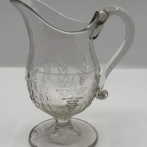 May include: Clear glass pitcher with a curved handle and a spout. The pitcher features etched leaf and grape designs around the body. It has a round base and a pedestal foot.