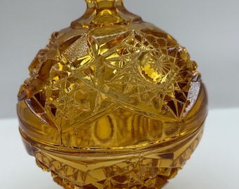 May include: Amber-colored, vintage glass candy dish with a lid. The dish has a round, textured body with a geometric pattern and sits on a pedestal base. The lid is topped with a diamond-shaped finial.