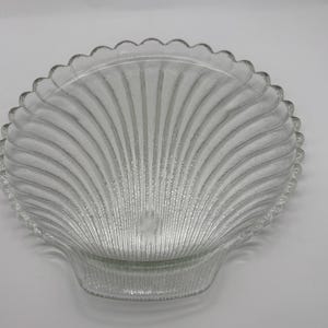 May include: A clear glass serving dish shaped like a seashell. The dish has a textured surface and a raised rim.