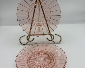 Puede incluir: Dos platos de cristal rosa con diseño radial, uno expuesto sobre un soporte metálico dorado. Los platos tienen un patrón texturizado en forma de abanico en los bordes. El soporte tiene un diseño curvo.