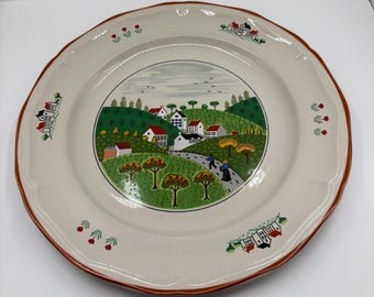 May include: Cream-colored ceramic plate with a village scene. The plate features a brown rim and a central illustration of houses, trees, and a road. Decorative floral and house motifs are placed around the rim. A charming piece for display or dining.