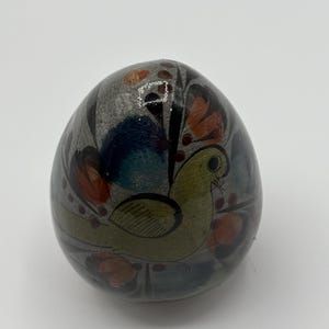 May include: A hand-painted ceramic egg with a bird design. The egg is decorated with blue, green, yellow, orange, and black paint.