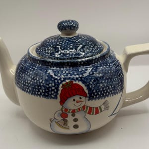 May include: A ceramic teapot with a white base and a blue speckled pattern on the lid and upper body. A snowman graphic with a red hat and scarf decorates the front. The teapot has a handle and spout.