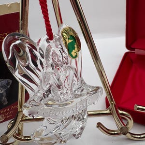 Op de afbeelding: Transparante glazen engel kerstornament met een rood koord om op te hangen. Het ornament heeft het woord "Marvstorp" erin geëtst. Een groen en goudkleurig label met een zeepaardje is bevestigd. Het ornament wordt op een gouden standaard getoond.