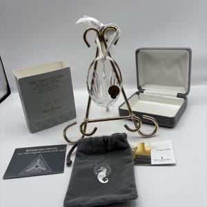 Peut inclure: Ornement en cristal transparent de la collection Waterford Times Square 2002, présenté sur un support en métal doré. Comprend une pochette en velours gris avec un emblème de hippocampe, une boîte grise et des cartes d'information.