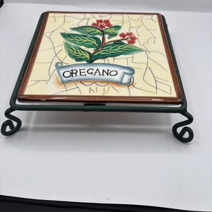 Könnte beinhalten: Ein dekorativer Keramik-Topfuntersetzer mit einem Oregano-Pflanzen-Design mit grünen Blättern und roten Beeren. Der Untersetzer hat einen cremefarbenen Hintergrund, einen braunen Rand und wird von einem grünen Metallständer getragen. Das Wort "OREGANO" ist auf einem Banner abgebildet.