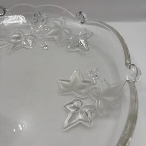 Puede incluir: Fuente de vidrio transparente con un diseño de hojas de hiedra en relieve. La fuente tiene un borde festoneado y dos asas pequeñas. Las hojas están dispersas por la superficie de la fuente.