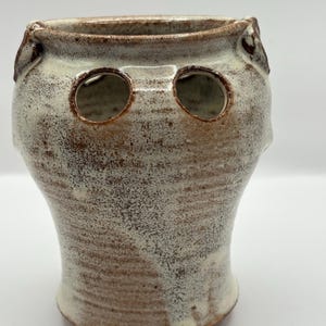 May include: A handmade ceramic vase with a unique design. The vase features a speckled brown and cream color scheme, with two circular openings near the top. The vase has a rounded body and a flared base, perfect for displaying flowers or as a decorative piece.