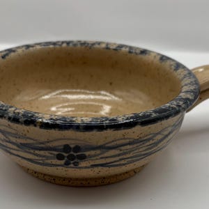 May include: A small, beige ceramic bowl with a blue and white floral design and a handle. The bowl is speckled with brown flecks.