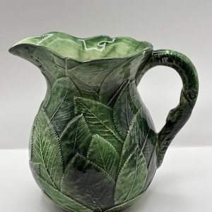 May include: A green ceramic pitcher with a leaf design. The pitcher has a wide spout and a curved handle. The body of the pitcher is covered in overlapping leaves, creating a textured surface. The pitcher is a dark green color.