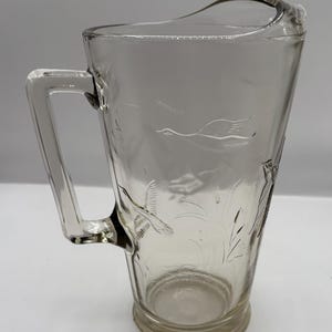 May include: A clear glass pitcher with a handle, featuring an etched design of birds and reeds. The pitcher has a slight chip on the rim.