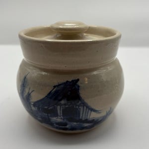 May include: A small, beige ceramic jar with a matching lid. The jar features a hand-painted blue and white illustration of a building. The jar has a rounded shape and a small handle on the lid.