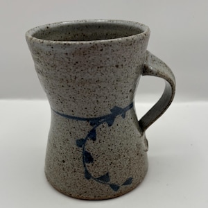 Puede incluir: Una taza de cerámica gris con un diseño floral azul y blanco. La taza tiene una base ancha y una parte superior estrecha.