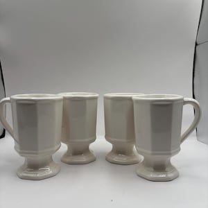 May include: Four off-white, footed mugs with a faceted design. Each mug has a handle and a pedestal base. The mugs are arranged in a row against a plain background. The mugs are made of ceramic.