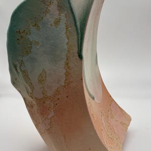 May include: A ceramic vase with a unique, abstract shape. The vase is glazed in shades of green, pink, and white, with a textured surface.