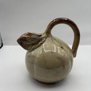 Puede incluir: Jarra de cerámica con cuerpo bulboso y asa curva. La jarra es de color beige moteado con detalles marrones en el asa y el pico. Un detalle en forma de hoja adorna el pico.