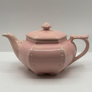 May include: A light pink ceramic teapot featuring a hexagonal body with a matching lid. The teapot has a curved spout, a sturdy handle, and decorative vertical ribbed detailing along the edges of the hexagonal panels and the lid.