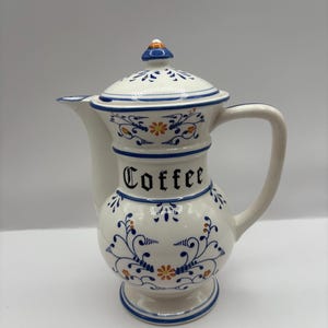 May include: White ceramic coffee pot with a lid, handle, and spout. The pot is decorated with blue floral patterns and the word "Coffee" in black script. Orange and yellow accents are also present.
