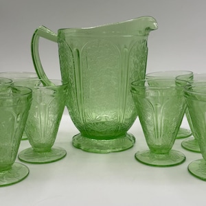 May include: A light green glass pitcher with a matching set of six footed glasses. Each piece features a detailed embossed floral pattern across the surface. The pitcher has a curved handle and a pouring spout, while the glasses have a tapered shape.