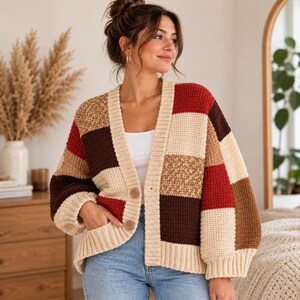 May include: A chunky crochet cardigan featuring a color-blocked patchwork design in shades of cream, dark brown, rust red, and tan. The garment has a V-neckline, long sleeves with ribbed cuffs, a ribbed hem, and two large wooden buttons on the front.