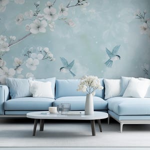 Puede incluir: Un sofá seccional azul claro con almohadas blancas se encuentra frente a un mural de pared con flores blancas y pájaros azules sobre un fondo azul claro.