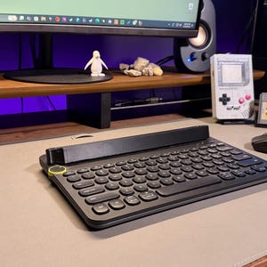 May include: A black wireless keyboard with a yellow accent sits on a gray desk mat. A black mouse, a decorative Game Boy, and a video game cartridge are also on the desk. A computer monitor and speakers are in the background.