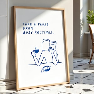 May include: A framed art print with a minimalist blue line drawing of a person reading a book and drinking coffee, with a croissant. The text reads "TAKE A PAUSE FROM BUSY ROUTINES." The frame is light brown, and the background is a room with a window.