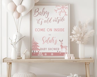 May include: A framed baby shower sign with the text "Baby it's cold outside, come on inside to Selah's baby shower." The sign features a pink and white winter theme with a tree, snowflakes, and a reindeer. The sign is displayed on a wooden table.