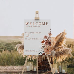 Pu&ograve; includere: Un cartello di benvenuto per matrimoni su un cavalletto. Il cartello recita "WELCOME TO OUR WEDDING" con i nomi William Thorn e Tessa Smithfield. Composizioni floreali ed erba della pampa decorano il cartello. Il cartello &egrave; all'aperto.