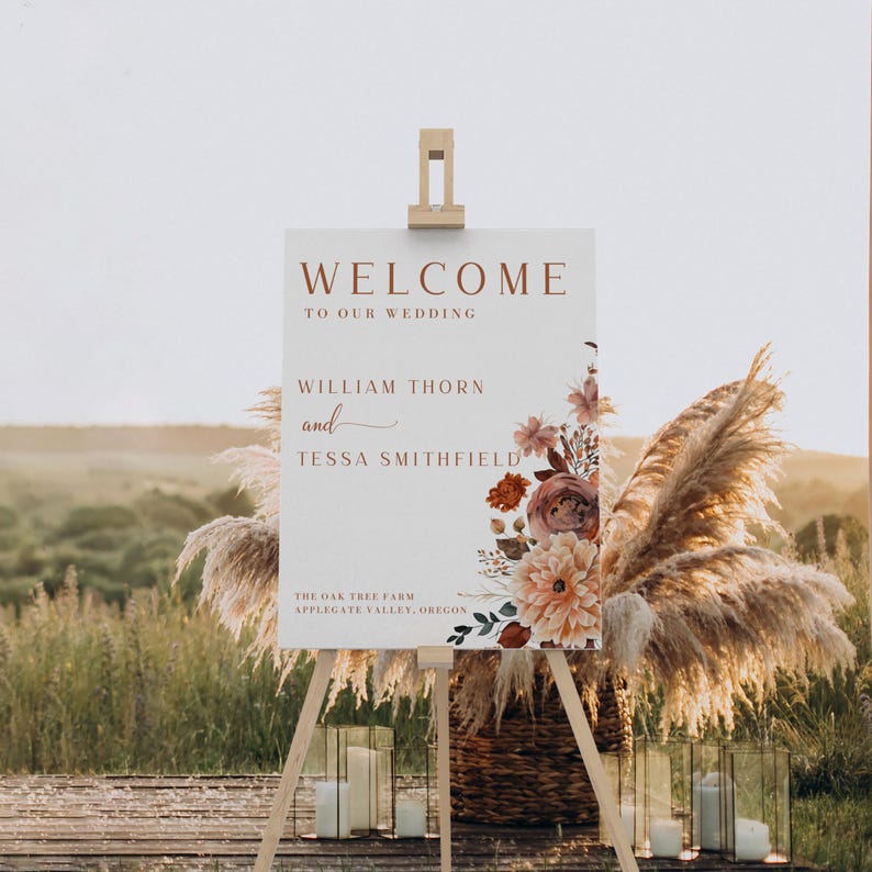 Pu&ograve; includere: Un cartello di benvenuto per matrimoni su un cavalletto. Il cartello recita "WELCOME TO OUR WEDDING" con i nomi William Thorn e Tessa Smithfield. Composizioni floreali ed erba della pampa decorano il cartello. Il cartello &egrave; all'aperto.