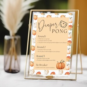 Puede incluir: Un letrero enmarcado con las palabras "Diaper Pong" en escritura elegante. El letrero presenta un tema de calabaza con calabazas naranjas y blancas. Las reglas del juego se enumeran debajo del título, perfecto para un baby shower.