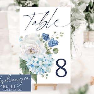 May include: White and blue floral table number card with the text "Table 8" in navy blue. The card is decorated with white peonies and blue hydrangeas. The text "Hydrangea Bliss Collection" is in the bottom left corner.