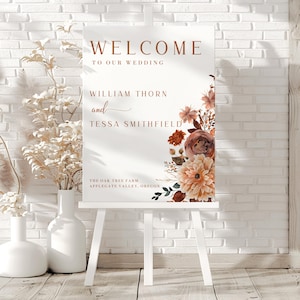 Pu&ograve; includere: Un cavalletto bianco espone un cartello di benvenuto per un matrimonio. Il cartello presenta le parole "WELCOME TO OUR WEDDING" in un carattere marrone caldo. Sotto, sono elencati nomi e un luogo, accompagnati da illustrazioni floreali nei toni del pesca e del marrone.
