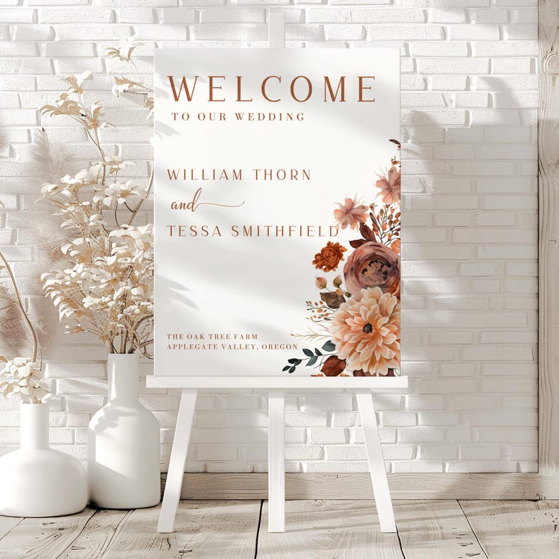 Pu&ograve; includere: Un cavalletto bianco espone un cartello di benvenuto per un matrimonio. Il cartello presenta le parole "WELCOME TO OUR WEDDING" in un carattere marrone caldo. Sotto, sono elencati nomi e un luogo, accompagnati da illustrazioni floreali nei toni del pesca e del marrone.