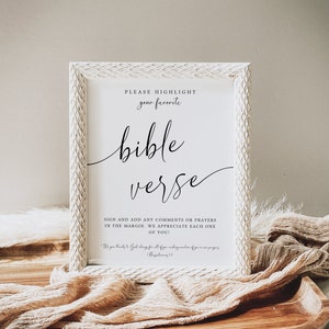 May include: White framed sign with the text "PLEASE HIGHLIGHT your favorite bible verse" and "SIGN AND ADD ANY COMMENTS OR PRAYERS IN THE MARGIN. WE APPRECIATE EACH ONE OF YOU!"  The sign is sitting on a wooden surface with a beige fabric draped over it.