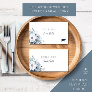 May include: Two white place cards with blue floral designs and the text "Table One Sarah Smith" and "Table Two Sarah Smith". The cards are on a wooden plate with a fork and knife on a white wooden surface.