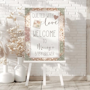 May include: A baby shower sign on a white easel. The sign reads "Quilted with love, Welcome to Maisy's Baby Shower." The sign has a floral and patchwork design with a heart and a small tag illustration.