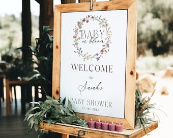 May include: A white sign with a floral wreath and the text "Baby in Bloom" welcomes guests to a baby shower for "Sairah's" on 3.16.2024. The sign is on a wooden easel.