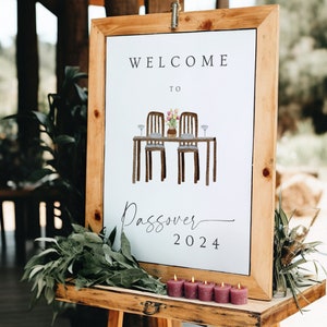May include: A wooden framed sign with the words "WELCOME TO" at the top and "Passover 2024" at the bottom. The sign features an illustration of a table set for two with flowers and glasses. Six small, purple candles are in front of the sign.
