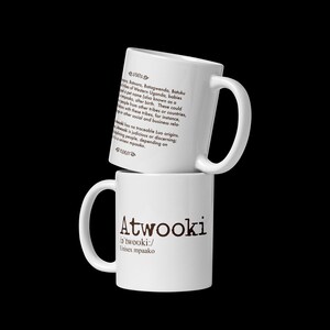 Puede incluir: Dos tazas de cerámica blancas apiladas. La taza superior tiene texto sobre los orígenes del nombre "Atwooki". La taza inferior muestra el nombre "Atwooki" en una fuente marrón oscuro, con pronunciación fonética y "Unisex mpaako".