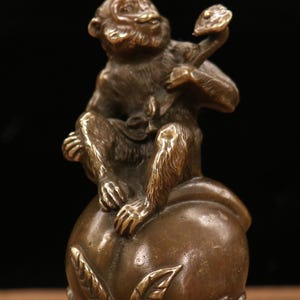 May include: Bronze sculpture of a monkey seated atop a large peach. The monkey is holding a smaller peach in its hand. The sculpture has a detailed, textured surface and is set against a dark background.