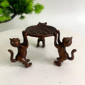May include: A bronze-colored cat figurine supporting a round, ornate tray. Two cats stand on their hind legs, lifting the tray, while a third cat supports it in the middle. The figurine is set against a white background.