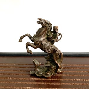 May include: A bronze sculpture of a monkey riding a horse. The horse is rearing up on its hind legs and the monkey is sitting on its back. The sculpture is set on a base of stylized waves.