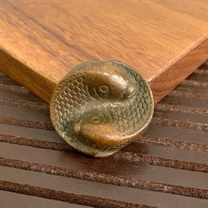 Può includere: Un oggetto circolare color bronzo con due pesci intrecciati in un design yin-yang. I pesci hanno squame dettagliate e sono posti su uno sfondo di legno. L'oggetto sembra essere un elemento decorativo o una piccola scultura.
