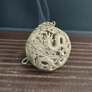 May include: A silver-colored, spherical incense burner with intricate dragon designs. The burner is open, and smoke is rising from within. The sphere is suspended by a chain and has a hinged opening.