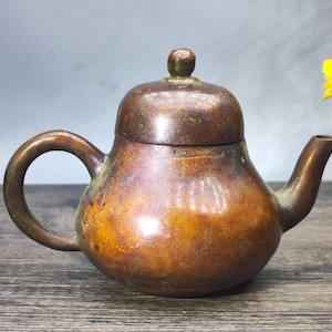 May include: A small, antique bronze teapot with a rounded body, curved handle, and a spout. The lid has a small knob. The teapot has a weathered appearance with variations in brown and gold tones, suggesting age and use. The teapot is likely used for brewing tea.